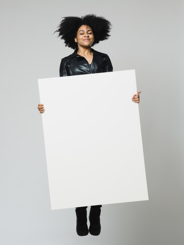 Full length portrait of cheerful young african woman jumping with eyes closed and holding a blank billboard. Vertical shot of real black woman with empty poster over gray background. Studio photography from a DSLR camera. Sharp focus on eyes.