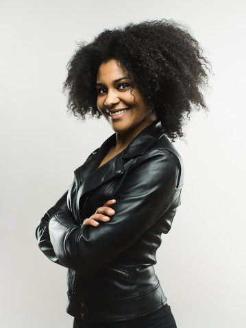 Portrait of smiling african woman looking at camera with her arms crossed against gray background. Vertical shot of stylish real woman in leather jacket. Studio photography from a DSLR camera. Sharp focus on eyes.