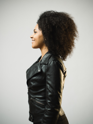 Portrait of smiling african woman looking to the side standing against gray background. Vertical shot of stylish real woman in leather jacket. Studio photography from a DSLR camera. Sharp focus on eyes.