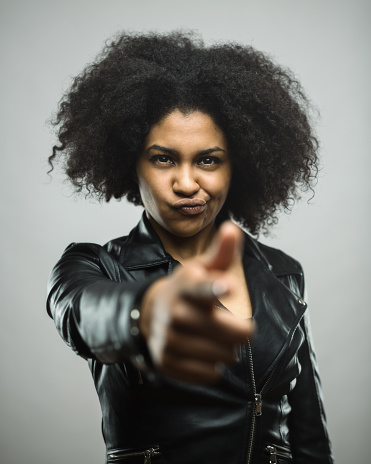 Vertical shot of angry young woman pointing at you against gray background. Real black woman portrait shooting with hand gun. Studio photography from a DSLR camera. Sharp focus on eyes.