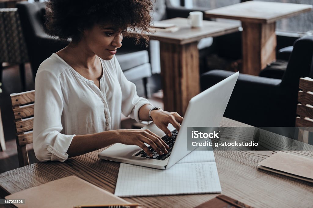 Developing new project. Beautiful young African woman using computer and smiling while sitting in cafe Adult Stock Photo