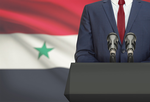 Businessman or politician making speech from behind the pulpit with national flag on background - Syria