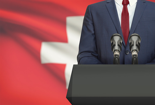 Businessman or politician making speech from behind the pulpit with national flag on background - Switzerland