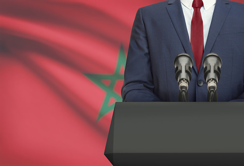 Businessman or politician making speech from behind the pulpit with national flag on background - Morocco