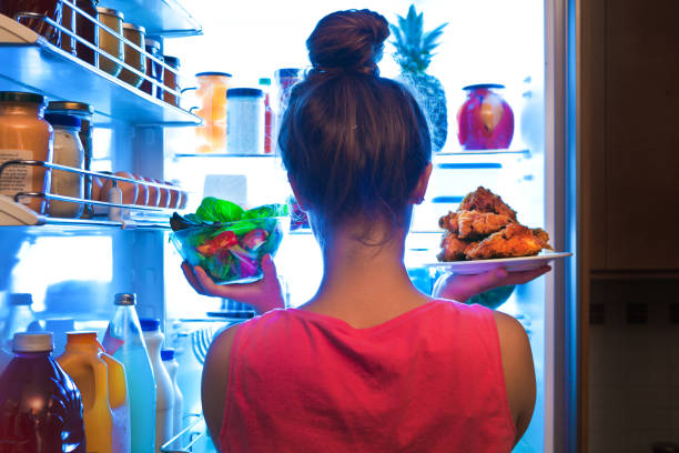 Young Woman Making Choices for a Healthy Salad or Junk Food Fried Chicken A young woman standing in front of the refrigerator, holding a bowl of fresh vegetable salad for a healthy diet on one hand and holding a plate of unhealthy fried chicken on the other. Making decision and choices for lifestyle and eating habit. unhealthy eating stock pictures, royalty-free photos & images