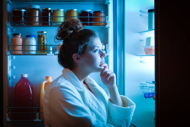 Dieting Young Woman Late Night Making Choices on What to Eat A young dieting woman standing in front of the refrigerator, contemplating and thinking about what to eat for hunger. Making choices and decision for healthy lifestyle hungry stock pictures, royalty-free photos & images