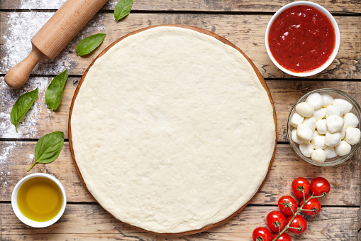 Raw dough preparation or pizza with ingredient: tomato sauce, mozzarella, tomatoes, basil, olive oil, spices served on rustic wooden table. Flat lay style, copy space for text. Margherita