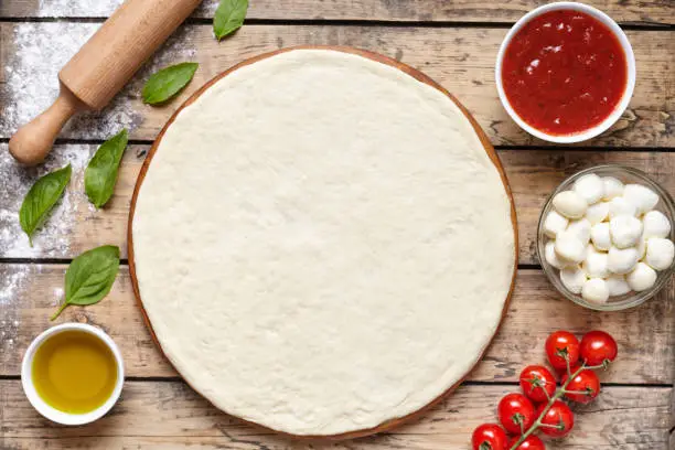 Photo of Raw dough for pizza with ingredient: tomato sauce, dough, mozzarella, tomatoes, basil, olive oil, spices served on rustic wooden table. Aerial shot, copyspace for text.