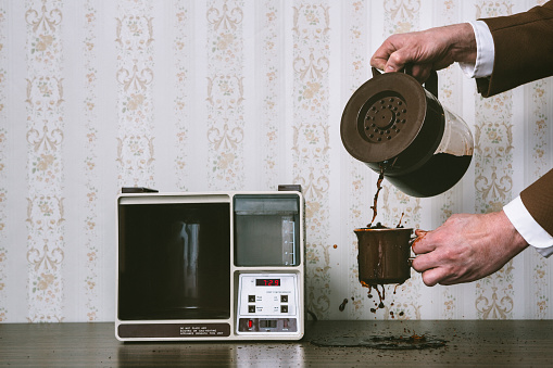 A man pours a cup of coffee from a vintage looking automatic coffee machine, spilling coffee all over.  The coffee maker sits on the counter of a 1970's / 1980's style kitchen with floral wallpaper.  Brown and yellow color theme.   Horizontal with copy space.