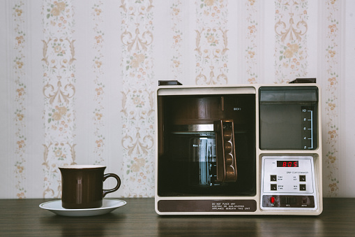 A vintage looking automatic coffee machine sits on the counter of a 1970's / 1980's style kitchen with floral wallpaper and a mug ready to be filled.  Brown and yellow color theme.   Horizontal with copy space.