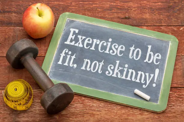 Exercise to be fit, not skinny! Fitness concept on a  slate blackboard against weathered red painted barn wood with a dumbbell, apple and tape measure