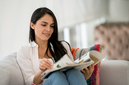 Happy woman relaxing at home sitting on the sofa reading a magazine - lifestyle concepts