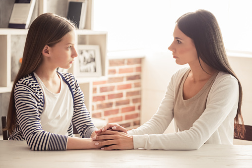 Troubled teenage girl and her mom are looking at each other and talking while sitting at home