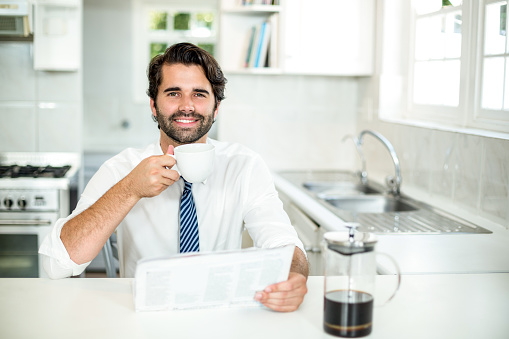 Portrait of confident businessman drinking coffee while reading newspaper in kitchen