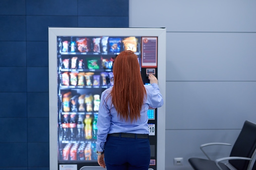 rear view of business woman inside airport sitting in front of vending machine and buying some snack.lifestyle shot.