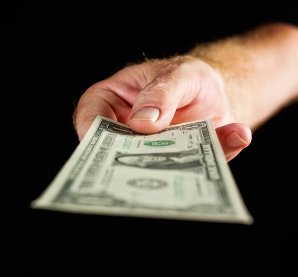 istock Male hand offers US one dollar banknote against black 667360098