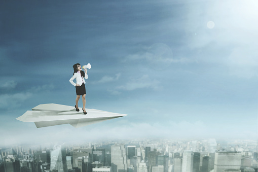 Female entrepreneur standing on a paper plane and yelling through a megaphone while flying above a city