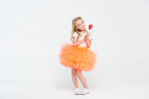 Cute little girl in festive dress holding heart shaped lollipop on white