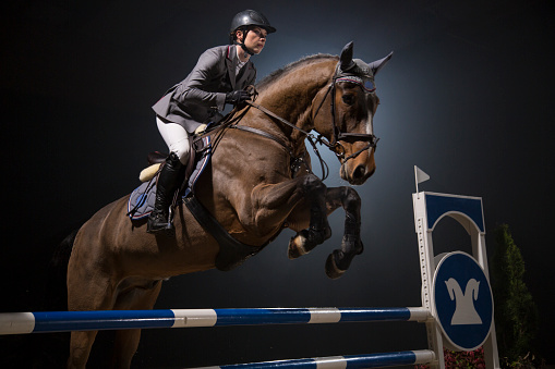 Female horseback rider jumping over hurdle at night.