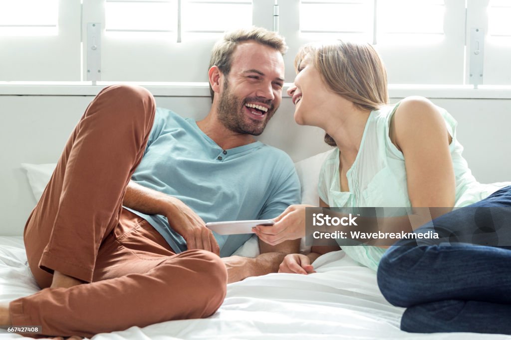 Couple with mobile phone laughing while relaxing on bed Couple with mobile phone laughing while relaxing on bed at home 30-34 Years Stock Photo