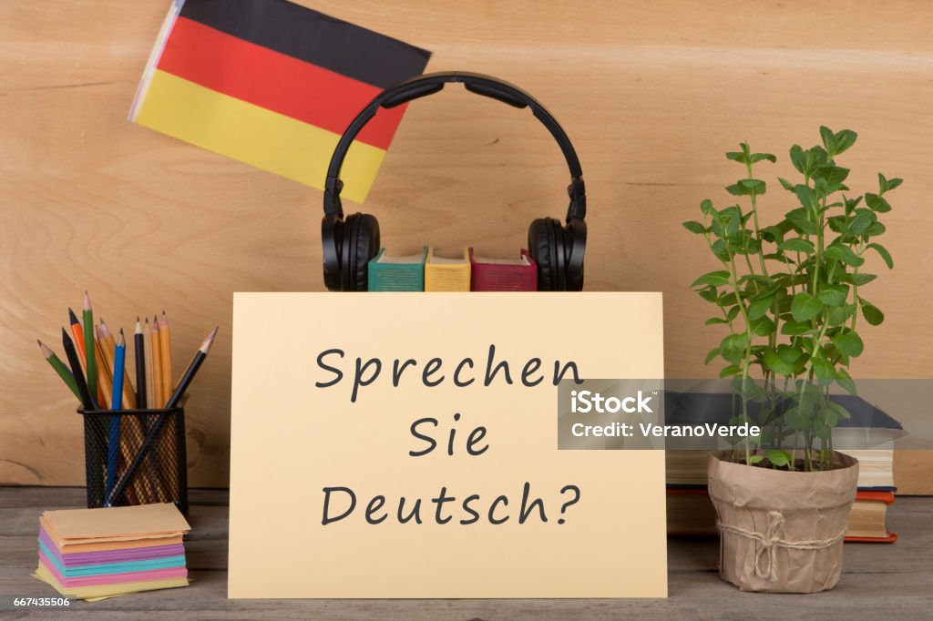 paper with text "sprechen sie deutsch?", flag of the Germany Learning languages concept - paper with text "sprechen sie deutsch?", flag of the Germany, books, headphones, pencils on wooden background German Language Stock Photo