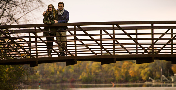 A teenage couple stands outside together on a bridge over looking a small river with colourful trees in the background and the front tires of a bike off to the left.
