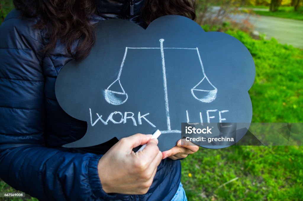 Work life balance dilemma concept with written words woman hands holding a blackboard with speech bubble shape with the words work and life. A drawing of a balance in chalk illustrates a difficult choice, comparison concept, and compromise. Life Balance Stock Photo