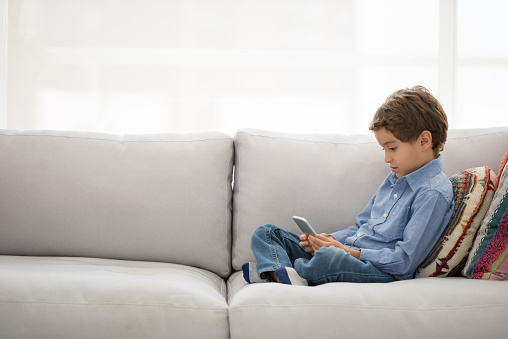 Beautiful little boy at home sitting on the sofa and using a smart phone at home