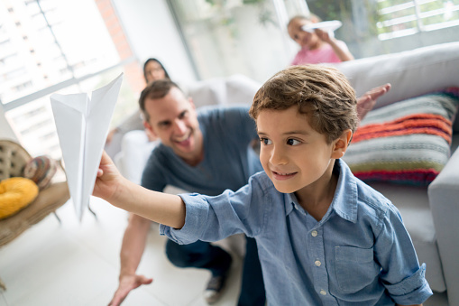 Portrait of a happy little boy playing with a paper plane at home and with his family at the background