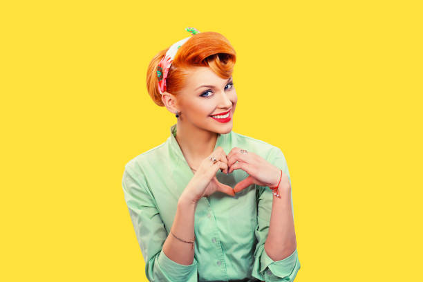 Love. Closeup portrait smiling happy pin up woman making heart sign symbol with hands isolated yellow wall background. Positive human emotion expression feeling life perception attitude body language stock photo