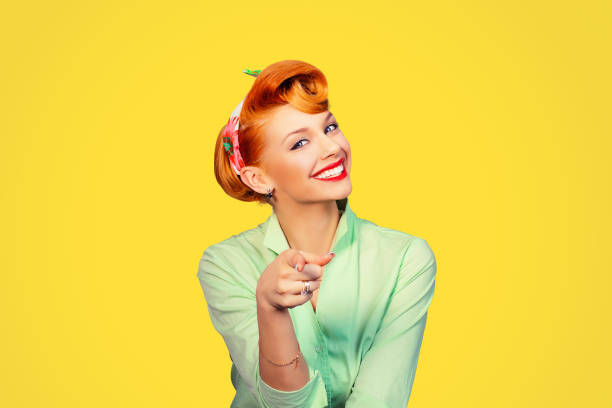 portrait of a beautiful woman pinup retro style pointing at you smiling laughing isolated yellow background wall. Body language, gestures, psychology. stock photo