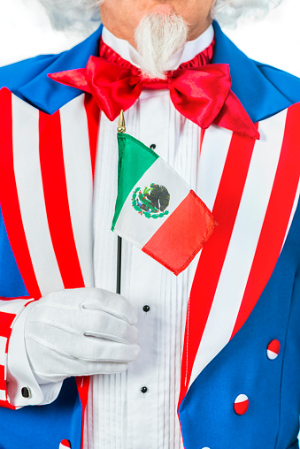 A close-up of a patriotic Uncle Sam character's white gloved hand holding a small Mexico flag in front of his chest. He is wearing a red, white and blue coat along with formal gloves.