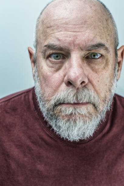 Too close-up for comfort creepy headshot portrait of a spooky, motionless 66 year old green eyed senior adult man staring suspiciously, impassively, and ominously at the camera. The skin on his face and balding head is showing his age, with scattered wrinkles, age spots, and blemishes. His remaining head hair is cut very short, and he has a full salt and pepper gray beard and mustache.