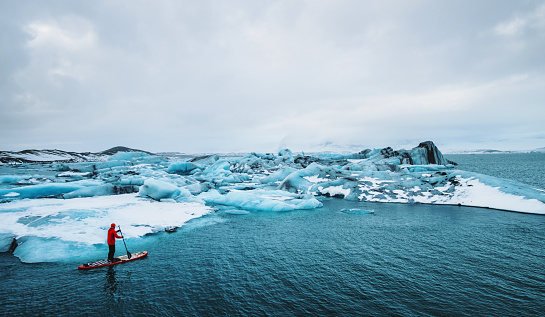 Beautiful view of icebergs glacier lagoon with a guy paddle boarding (sup), global warming and climate change concept