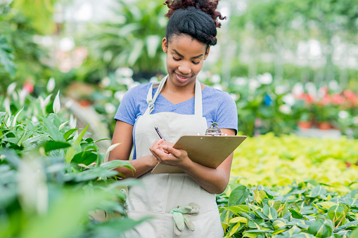 An African American woman is inside a plant nursery. She is wearing gardening clothes. She is facing smiling while using a clipboard.
