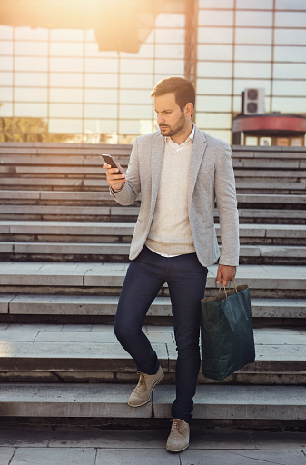 Young man in smart casual clothing reading text message on cell phone while carrying shopping bag and going down the stairs in the city.