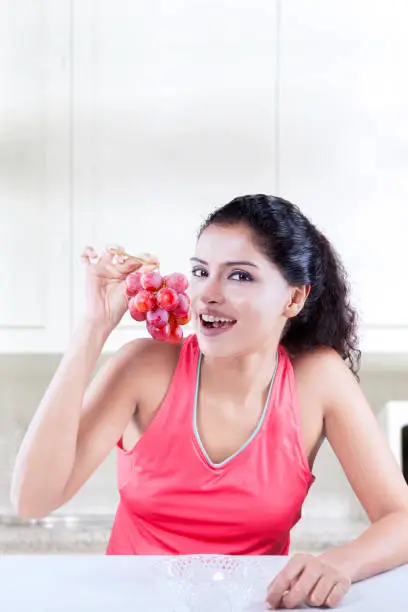 Indian woman looking at the camera while sitting in the kitchen with a bunch of fresh grapes