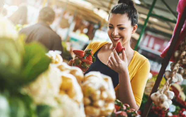 Woman buying fresh fruits and vegetables. Closeup front view of late 20's attractive woman choosing some fruit at local food market.She's holding a pack of strawberries and smiling. She's wearing a nice casual yellow blouse. fruit cart vendor stock pictures, royalty-free photos & images