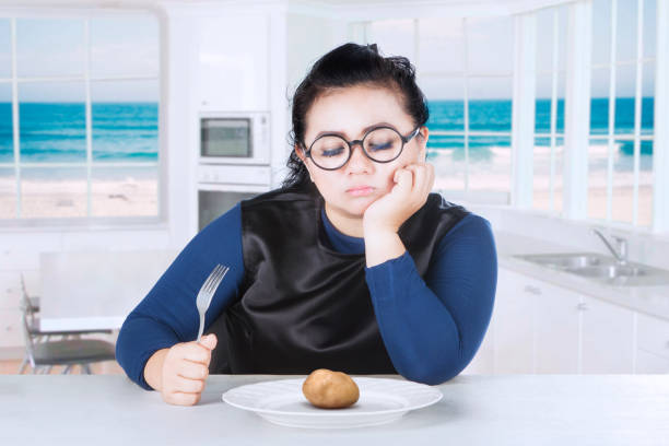 Fat woman feeling bored with boiled potato while holding a fork, beach background on the window