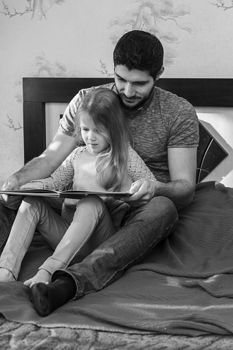 Dad and daughter read a book. They sit on the bed in the room.