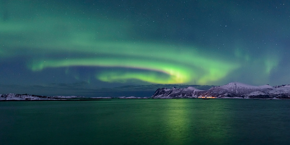 Northern Lights, polar light or Aurora Borealis in the night sky over Senja island in Northern Norway. Snow covered mountains in the background with water of the Norwegian Sea reflecting the lights in the foreground.