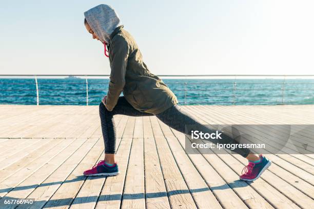 Woman In Jacket And Hood Doing Sports Exercises On The Beach In The Morning Stock Photo - Download Image Now
