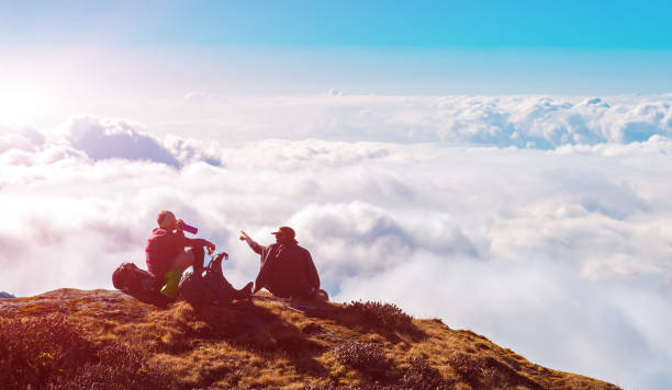 istock Two People sitting on high Cliff above clouds in Mountains 667770506