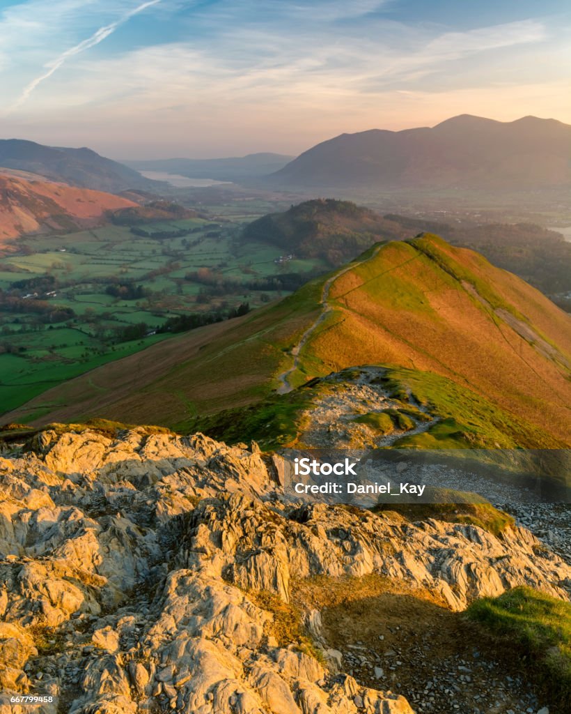 Catbells in the Lake District at sunrise. Vibrant sunrise colours with golden light on rocks in the foreground taken in the English Lake District on a spring morning. Awe Stock Photo