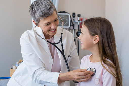 Happy little girl at the doctor for a checkup. Friendly pediatrician examining girl with stethoscope in her office. Happy senior woman auscultate the heartbeat of the child.