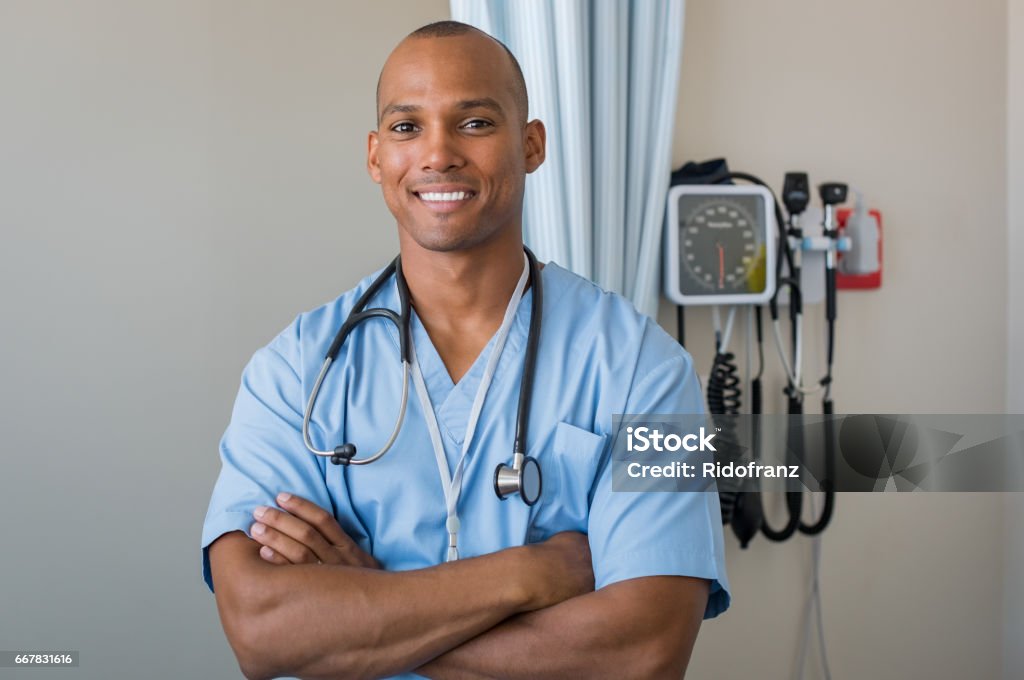 Happy nurse smiling Cheerful african doctor standing with crossed arms in hospital. Pride male nurse looking at camera. Successful man surgeon wearing blue uniform with stethoscope around neck. Male Nurse Stock Photo