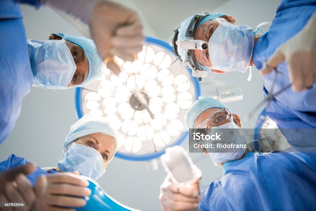Team of surgeons operating Surgeons team at work in operating room. Below view of surgeons holding medical instruments in hands and looking at patient. Multiethnic doctors wearing protective uniforms while operating. Emergency Room Stock Photo