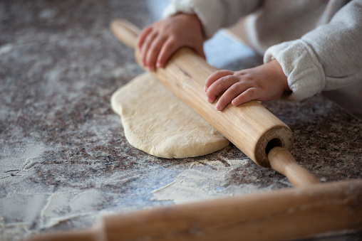 Little baker girl kneading dough with wooden rolling pin on marble table.