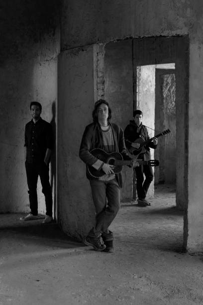 Young talents Black and white shot of a young musician with an acoustic guitar leaning by the wall while rock band members standing in the background. duo band photos stock pictures, royalty-free photos & images
