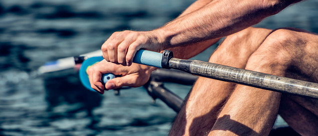 Close up of a rower’s hands holding oar and legs during rowing, panoramic.